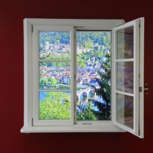 Künstliches Fenster ("Fake Window") mit echtem, weißem Fensterrahmen, geöffnetem Fensterflügel und Aussicht auf Heidelberg als Tageslichtfenster in einer roten Wand.